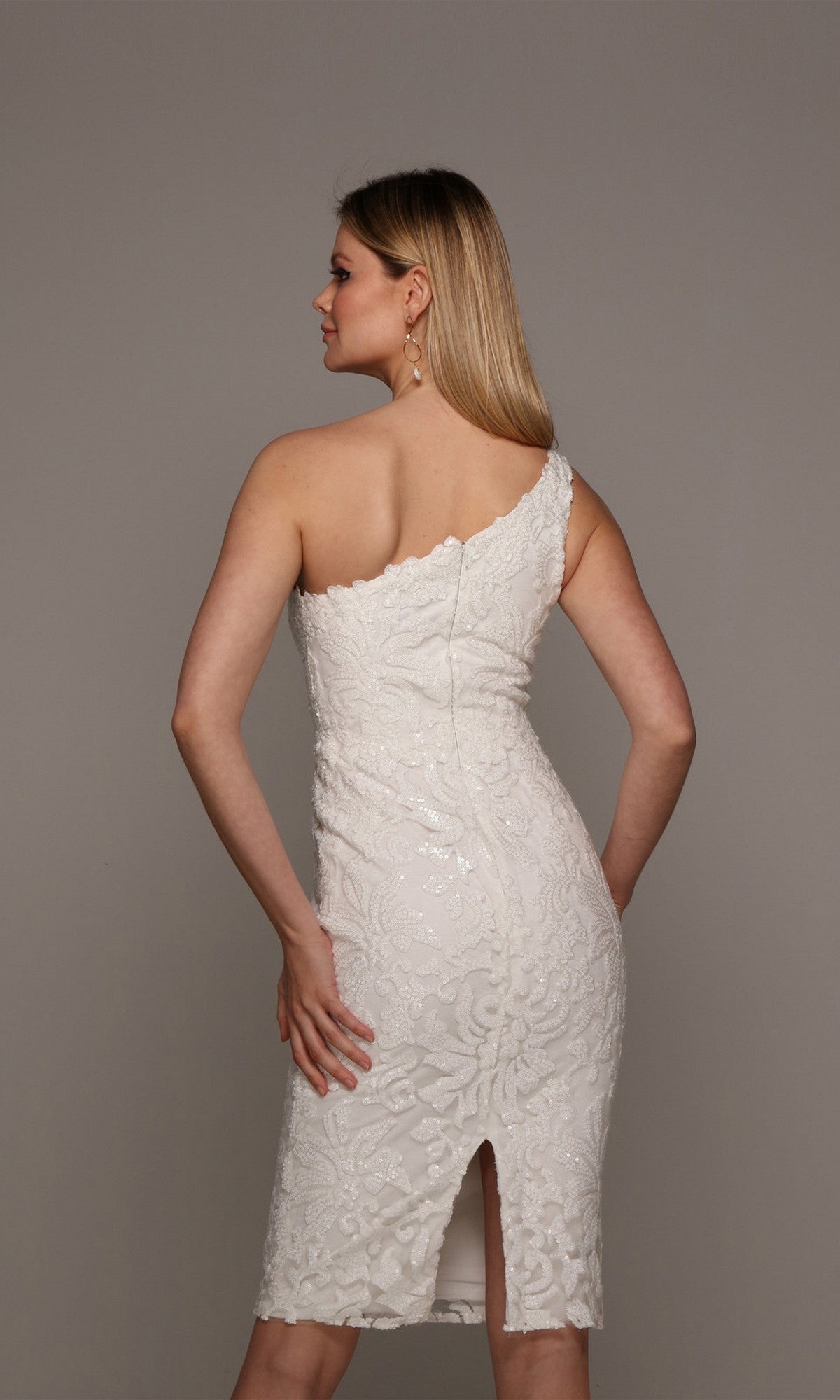 A woman with long blonde hair wears the Alyce 70029 knee-length white party dress featuring sequin lace, one-shoulder design, and a back slit, standing against a plain gray background.